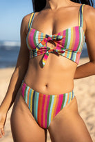 A woman modeling a vibrant colored vertically striped bralette bikini top with keyhole cutouts and double tie details paired with a high-waisted cheeky bikini bottom, photographed outdoors on a sandy beach with the ocean and blue sky in the background.