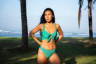Woman wearing a bright emerald green bralette bikini top and high-waisted cheeky bikini bottom outdoors with palm trees.