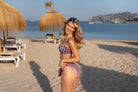 A woman posing on a sandy beach wearing a red, white, and blue vertically striped bikini featuring a removable strap bandeau and a scrunch butt ruched cheeky bikini bottom, with beach umbrellas, lounge chairs, and the blue sea in the background.
