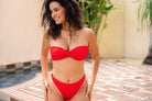 A woman modeling a bright red bikini with a bandeau-style top and high-cut, high-waisted cheeky bottoms, standing outdoors near a hotel tiled poolside area with a beige wall and green plants in the background.