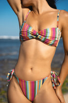 A woman wearing a colorful vertically striped bikini modeling a bandeau bikini top with removable straps and a string bikini bottom with tie-sides and a cheeky cut, photographed outdoors against a blurred beach and blue sky background.